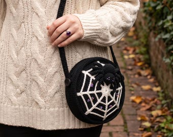 May include: A round black crochet crossbody bag featuring a white spiderweb design on the front. A three-dimensional black crocheted spider is attached to the web, accented with small purple beads embedded within the web pattern and on the spider.