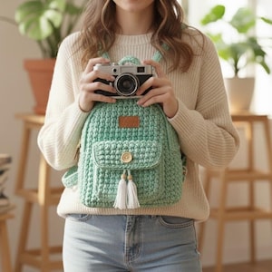 May include: A mint green crochet backpack with a front pocket, wooden button, and white tassels. The backpack has a small leather tag. A vintage camera is held in front of the backpack.