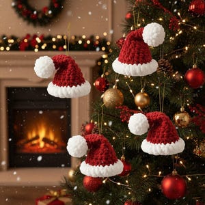 Może przedstawiać: Szydełkowe ozdoby świąteczne w kształcie czapek Świętego Mikołaja. Czapki są czerwone z białym obszyciem i białym pomponem. Wiszą na złotych sznurkach na udekorowanej choince.
