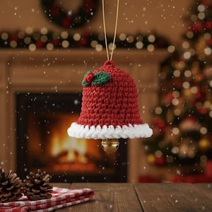 Puede incluir: Adorno navideño de campana roja de ganchillo con ribete blanco y una pequeña campana dorada. El adorno está decorado con una hoja verde y una baya roja, colgando de un hilo dorado.