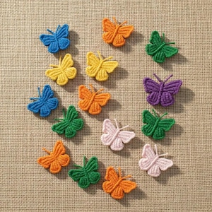Può includere: Una collezione di applicazioni a farfalla all'uncinetto colorate in varie tonalità di blu, arancione, verde, giallo, viola e rosa. Le farfalle sono disposte su uno sfondo neutro e strutturato, mettendo in risalto i loro dettagliati motivi alari e le antenne.