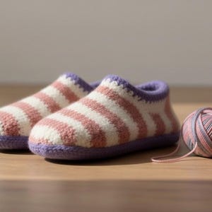 以下が含まれることがあります： ピンクと白のストライプ柄の手編みスリッパ。紫色のトリムが施されています。近くには、同系色の毛糸玉があります。スリッパは室内履き用で、柔らかい糸でできています。