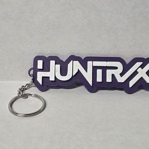 May include: A purple and white keychain with the word "HUNTRAX" in a bold, blocky font. The keychain has a silver key ring attached. The design is a simple, modern style.