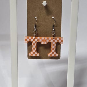 May include: A pair of orange and white checkered earrings in the shape of the letter "T". The earrings are hanging from silver-colored fish hook ear wires. The earrings are displayed on a brown card.