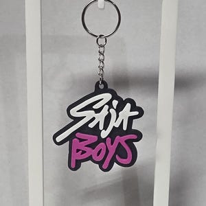 May include: Black and white keychain with the text "Skit Boys" in a stylized font, with pink accents. The keychain is attached to a silver metal ring and chain.