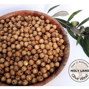 May include: A wooden bowl filled with numerous olive wood beads. The beads are light brown with natural wood grain patterns. Olive branches with green leaves and olives are on the right. A circular label reads "Made in Bethlehem, Holy Land, Olive Wood."