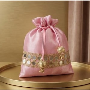May include: A light pink drawstring pouch made of a smooth, satin-like fabric. The bag features a horizontal band of gold-toned embroidery with circular mirror accents. Two matching pink cords with gold-toned, spherical, beaded tassels hang down.