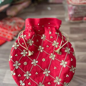 May include: A red drawstring pouch with gold floral embroidery and small pearl accents. The pouch has a round shape and is tied with a cream-colored cord. The background is blurred, suggesting a display of similar items.