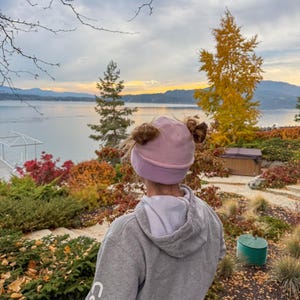 May include: A person wearing a light pink beanie with two bun-like features, facing a lake and colourful foliage. The person is wearing a grey hooded sweatshirt. The background includes a lake, trees, and a cloudy sky.