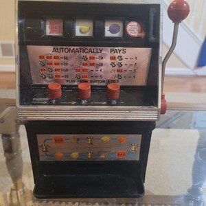 May include: A black and silver tabletop slot machine featuring three spinning reels with fruit and bar symbols. The front panel displays an 'AUTOMATICALLY PAYS' chart, three red push buttons labeled 1, 2, and 3, and a red-handled lever on the right side.