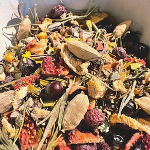 May include: Close-up of a mix of dried herbs and spices in a white bowl. The blend includes green, brown, red, and yellow components, with visible whole spices and dried berries. The arrangement suggests a culinary or herbal preparation.