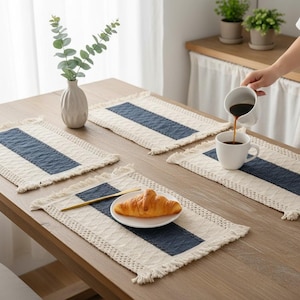 Op de afbeelding: Een houten tafel met vier rechthoekige placemats. Elke placemat heeft een crèmekleurige achtergrond met een marineblauwe streep in het midden en gefranjerde randen. Een witte vaas met eucalyptus, een bord met een croissant en een kopje koffie staan op tafel.