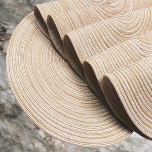 Puede incluir: Primer plano de un mantel individual tejido beige y blanco con un diseño circular. El mantel individual está parcialmente enrollado, revelando su construcción en capas. La textura parece estar hecha de fibras naturales, creando una estética neutra y terrosa.
