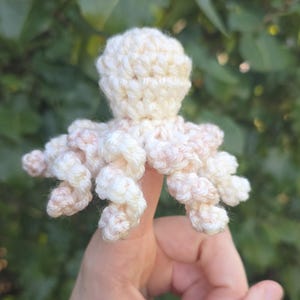 May include: A small, off-white crochet octopus toy featuring a rounded head and eight spiraled, curly tentacles. The yarn has subtle, light pinkish-peach color variations throughout the texture, creating a soft, multi-tonal appearance.