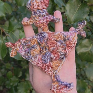 May include: A small, hand-crocheted ornament featuring a variegated yarn in shades of orange, purple, and cream. The piece has a central triangular shape with three distinct, pointed extensions radiating outward, creating a star-like or abstract form.