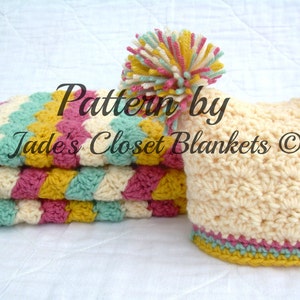 May include: A crocheted baby blanket in pink, yellow, cream, and green with a matching crocheted hat with a pom-pom. The blanket is folded in thirds and the hat is lying on its side. The text "Pattern by Jade's Closet Blankets ©" is visible on the blanket.