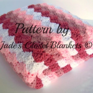 May include: A crocheted baby blanket in shades of pink, white, and red. The blanket is folded in half and has a textured pattern. The text "Pattern by Jade's Closet Blankets ©" is visible on the blanket.