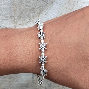 Peut inclure: Un bracelet de couleur argentée composé d'une série de petites breloques en forme de tortue détaillées. Chaque breloque tortue est séparée par une petite perle ronde de couleur argentée, créant un motif répétitif le long de la chaîne.