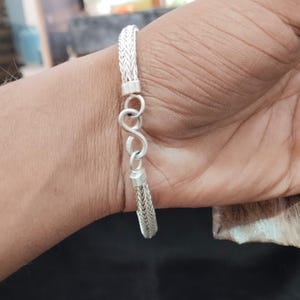 Peut inclure: Un bracelet en argent avec un motif de chaîne tressée et un symbole infini. Le bracelet est présenté sur un poignet, avec le fermoir visible. Le métal a une finition brillante et réfléchissante.