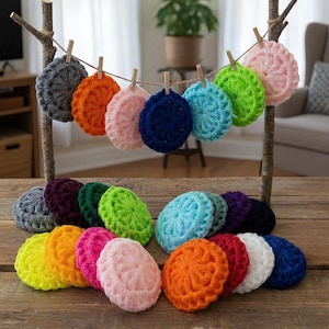May include: A collection of round, textured crochet scrubbies in a variety of vibrant colors including pink, orange, yellow, green, blue, purple, white, and dark gray. Some are hanging from a twine line on wooden branches, others are stacked.