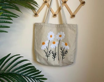May include: A beige canvas tote bag featuring a daisy floral design. The daisies have white petals and orange centres, with black stems and leaves. The bag is hanging on a wooden wall rack.