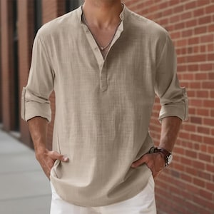 Peut inclure: Une chemise Henley beige à manches longues avec un col officier et une patte de boutonnage. Les manches sont retroussées. La chemise est en tissu léger. La personne porte une montre et un collier argenté.