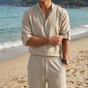 May include: Beige linen shirt with a mandarin collar and button-down placket. The long sleeves are rolled up. The shirt is paired with matching beige drawstring pants. A watch with a brown leather strap is visible.