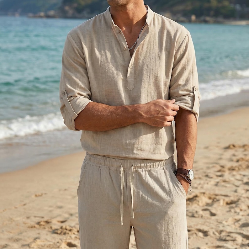 May include: Beige linen shirt with a mandarin collar and button-down placket. The long sleeves are rolled up. The shirt is paired with matching beige drawstring pants. A watch with a brown leather strap is visible.
