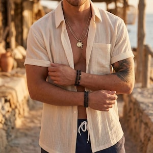 Può includere: Camicia color crema a maniche corte con maniche arrotolate. La persona indossa una collana d'oro con un ciondolo e due braccialetti di perline nere. La camicia è sbottonata, rivelando il petto.