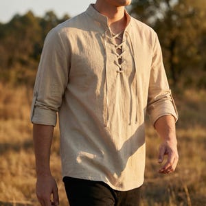 Puede incluir: Camisa de lino beige con cuello mandarín y cordones en la parte delantera. Las mangas largas están enrolladas. La camisa está hecha de una tela ligera, adecuada para climas cálidos. La camisa es de estilo casual.