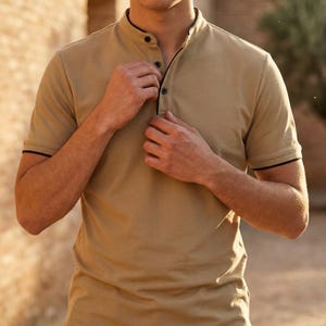 May include: Tan short-sleeved shirt with a mandarin collar and black trim. The shirt has three buttons down the front and is being buttoned up. The sleeves also have black trim.