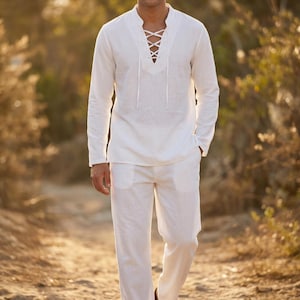 Può includere: Un completo camicia e pantaloni in lino bianco. La camicia a maniche lunghe presenta uno scollo allacciato. I pantaloni hanno tasche. L'outfit è adatto per il clima caldo e le occasioni informali.