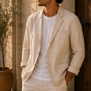 Puede incluir: Una americana de color beige con solapa de muesca, dos botones frontales y dos bolsillos de parche, combinada con una camiseta blanca lisa. La prenda presenta mangas largas con puños vueltos y un acabado de tela texturizada, de corte estructurado.