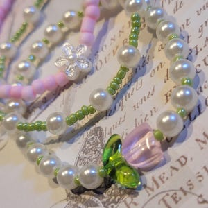 May include: Close-up of three beaded bracelets. One bracelet is pink with a silver flower charm. Another is white with green beads. The third bracelet has white beads, green beads, and a pink and green flower charm. The bracelets are on a vintage paper background.