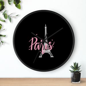 May include: Black round wall clock featuring the Eiffel Tower in white and the word "Paris" in pink script. Pink hearts are scattered around the design. The clock has black hands and a black frame.