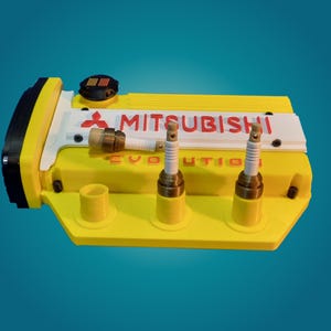 Puede incluir: Una cubierta de motor Mitsubishi Evolution amarilla y negra con la palabra "MITSUBISHI" en rojo. La cubierta tiene dos bujías y la palabra "EVOLUTION" en rojo. El fondo es un degradado de azul.