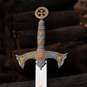 May include: An ornate sword with a silver blade and a detailed hilt. The hilt features gold accents, intricate engravings, and a cross-shaped emblem at the pommel. The sword rests against a backdrop of dark brown logs.