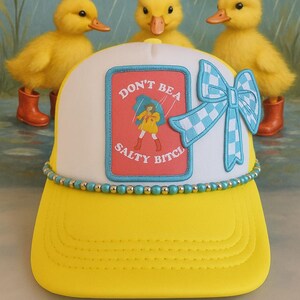 May include: A yellow and white trucker hat with the text "DON'T BE A SALTY BITCH" on a patch. The hat features a blue and white checkered bow and a beaded band. Three yellow ducklings in orange boots are in the background.