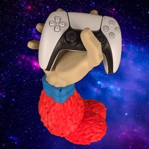 Pode incluir: Uma escultura de uma mão segurando um controle de videogame branco e preto. A mão é bronzeada, com uma manga azul e uma base vermelha texturizada. O fundo é uma galáxia colorida.