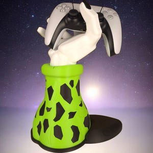 Peut inclure: Support de manette de jeu en forme de main blanche tenant une manette blanche et noire. La base verte en forme de vase est ornée de formes noires irrégulières. Le tout repose sur une plateforme circulaire noire. Fond de ciel étoilé.