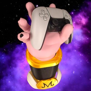 Può includere: Una scultura a forma di mano rosa che tiene in mano un controller di gioco bianco e nero. La scultura ha una base dorata e nera con un colletto bianco e un logo "M" giallo. Lo sfondo è una galassia viola e nera.