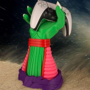 Op de afbeelding: Een groene handvormige gamecontrollerstandaard met een wit en zwarte controller. De standaard heeft een rode band, groene en roze secties en een paarse basis. De achtergrond is een sterrennacht.