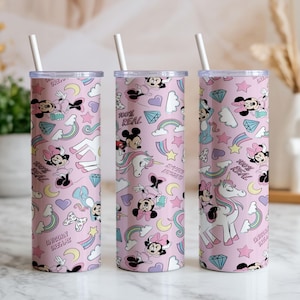 May include: Three pink insulated tumblers with white straws. The tumblers feature a repeating pattern of Minnie Mouse, unicorns, rainbows, stars, and diamonds. Text on the tumblers reads "100% REAL" and "UNICORN DREAM".