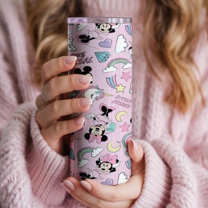 May include: A pink tumbler featuring a repeating pattern of cartoon characters, rainbows, stars, and the text "100% REAL". The tumbler has a silver lid and is held by a person wearing a pink sweater. The tumbler is cylindrical.