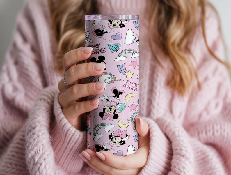 May include: A pink tumbler featuring a repeating pattern of cartoon characters, rainbows, stars, and the text "100% REAL". The tumbler has a silver lid and is held by a person wearing a pink sweater. The tumbler is cylindrical.