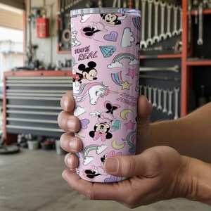 May include: A pink tumbler with a repeating pattern of Mickey and Minnie Mouse, unicorns, rainbows, and the text "100% REAL". The tumbler is cylindrical and appears to be made of stainless steel.