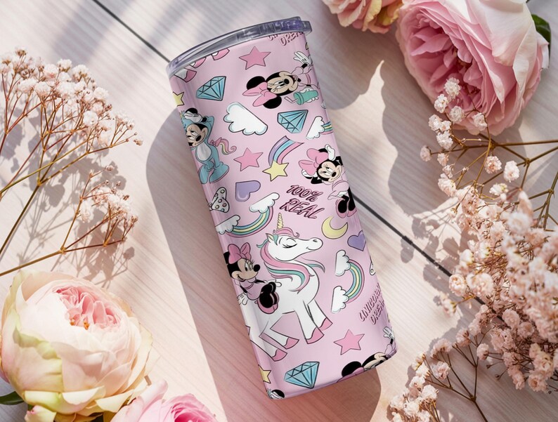 May include: A pink tumbler featuring a repeating pattern of Minnie Mouse, unicorns, rainbows, diamonds, and stars. The words "100% REAL" are printed on the tumbler. The tumbler is surrounded by pink roses and white flowers.