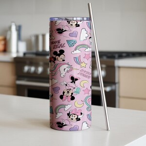 May include: A pink tumbler with a repeating pattern of cartoon characters, rainbows, stars, and the text "100% REAL". A silver-colored straw is next to the tumbler. The tumbler is cylindrical and appears to be made of metal.