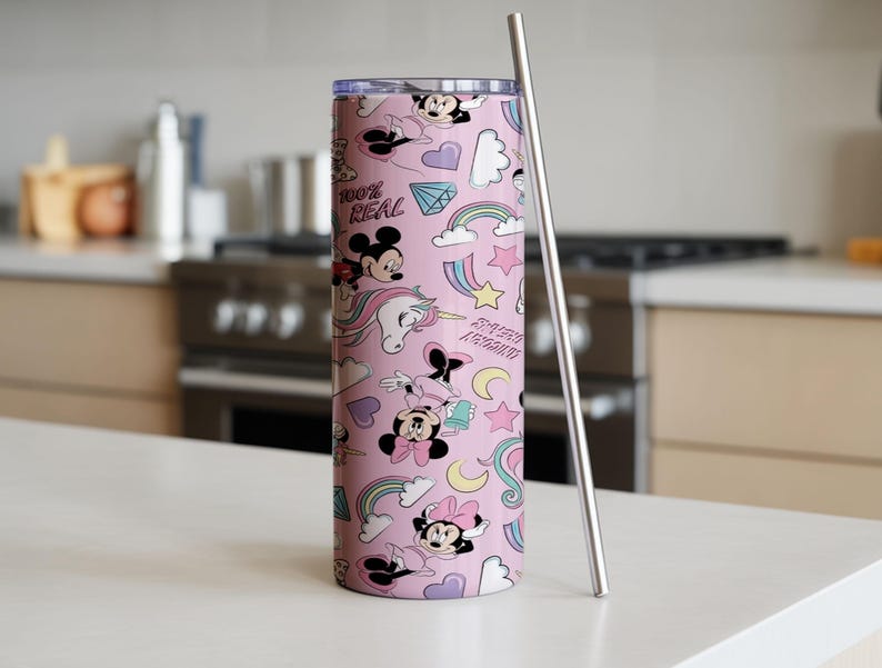 May include: A pink tumbler with a repeating pattern of cartoon characters, rainbows, stars, and the text "100% REAL". A silver-colored straw is next to the tumbler. The tumbler is cylindrical and appears to be made of metal.