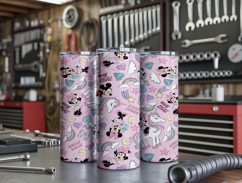 May include: Three pink insulated tumblers with a repeating pattern of Minnie Mouse, unicorns, rainbows, and diamonds. The tumblers have the text "100% REAL" and "UNICORN DREAMS". The tumblers are cylindrical and appear to be made of stainless steel.